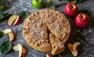 A homemade apple pie with a crumbly topping, sliced and served with fresh apples and apple slices on a dark surface.