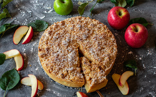 A homemade apple pie with a crumbly topping, sliced and served with fresh apples and apple slices on a dark surface.