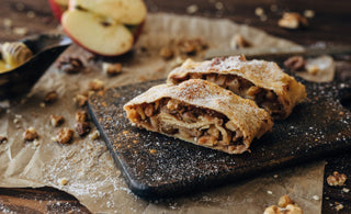 Two slices of apple strudel with powdered sugar on a dark cutting board, surrounded by walnuts, apple slices, and parchment paper.