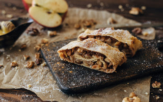 Two slices of apple strudel with powdered sugar on a dark cutting board, surrounded by walnuts, apple slices, and parchment paper.