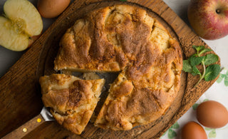Image of an apple cake with a slice removed, served on a wooden board, surrounded by apples, eggs, and mint leaves.