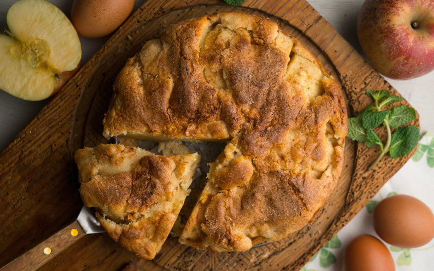 Image of an apple cake with a slice removed, served on a wooden board, surrounded by apples, eggs, and mint leaves.