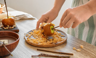 Hands dipping a candied apple in cereal on a wooden plate, with another caramel apple and bowl nearby on a wooden table.