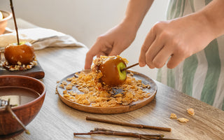 Hands dipping a candied apple in cereal on a wooden plate, with another caramel apple and bowl nearby on a wooden table.