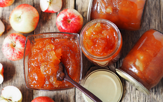 Bowl and jars of apple jam with a spoon, surrounded by red and sliced apples on a rustic wooden surface.