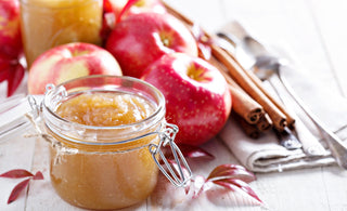 A glass jar of homemade apple butter or applesauce, surrounded by fresh red apples and cinnamon sticks on a rustic white wooden surface.
