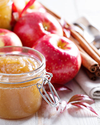 A glass jar of homemade apple butter or applesauce, surrounded by fresh red apples and cinnamon sticks on a rustic white wooden surface.