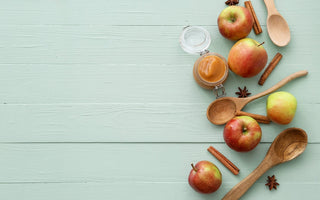 Overhead flat lay of a jar of apple butter, fresh apples, cinnamon sticks, star anise, and wooden spoons, arranged on a pale green wooden background with copy space.