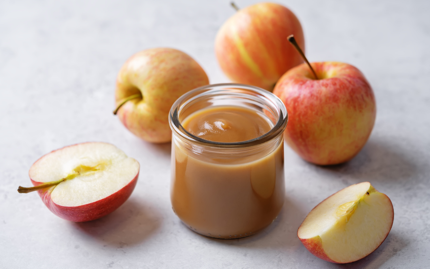 A jar of caramel sauce surrounded by whole and sliced apples on a light surface, highlighting the caramel sauce.