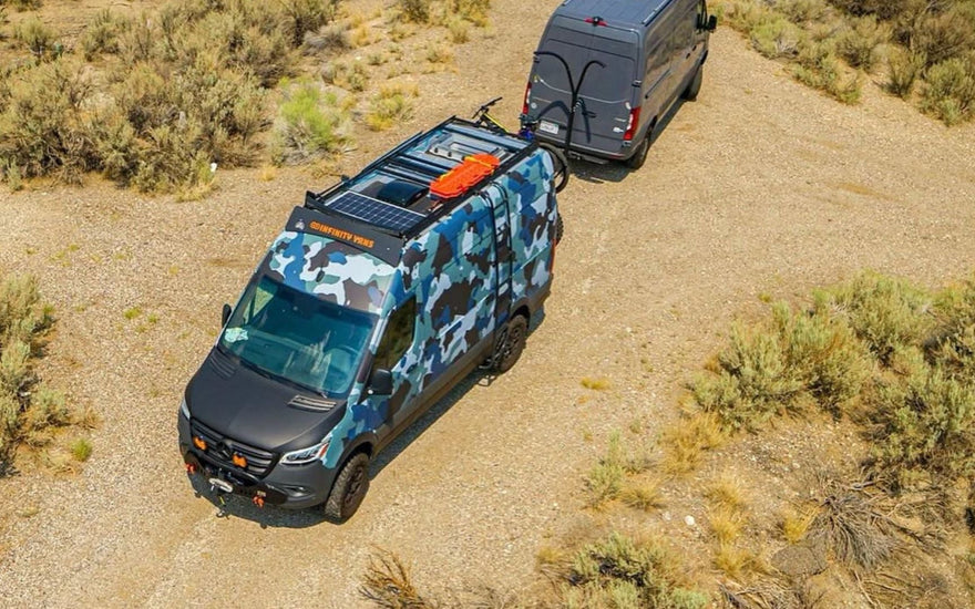  Aerial view of two Mercedes Sprinter vans parked on desert dirt road - one with blue camouflage wrap and solar panels, one grey - showcasing van life adventure travel