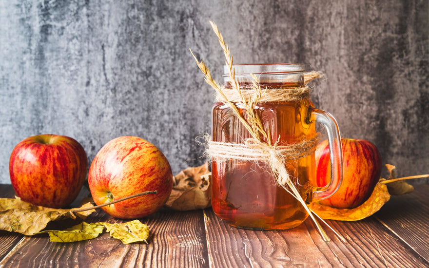 A mug filled with rich apple cider syrup wrapped in thick ropes for a rustic touch. Surrounding the mug are fresh apples and vibrant autumn leaves, all placed on a wooden surface, creating a warm and cozy fall atmosphere.