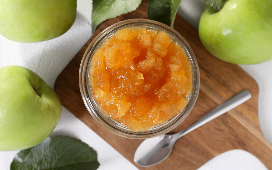 Jar of apple jam with spoon on wooden board, surrounded by green apples and leaves.